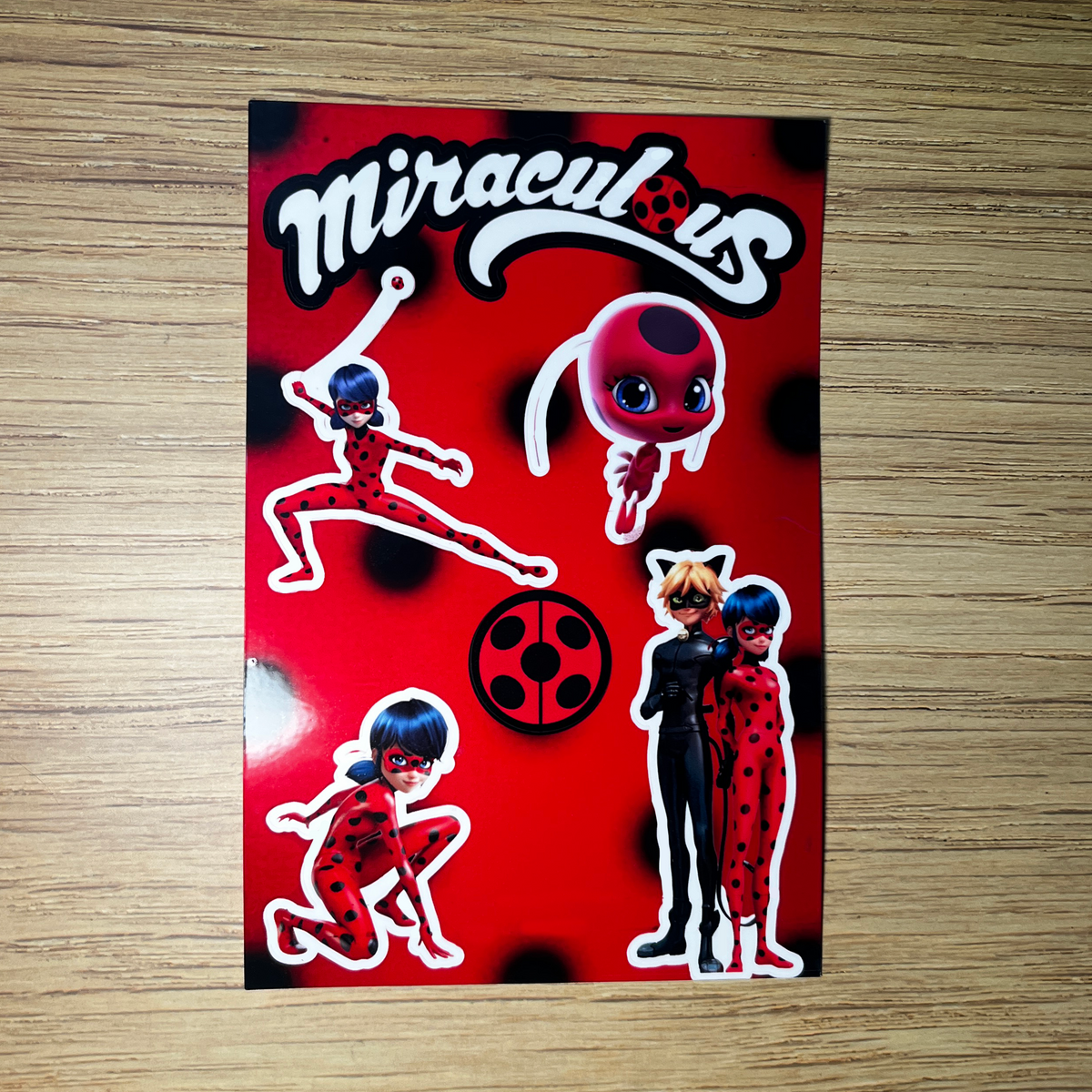 Sticker with 'Miraculous' characters on a red background, placed on a wooden surface.