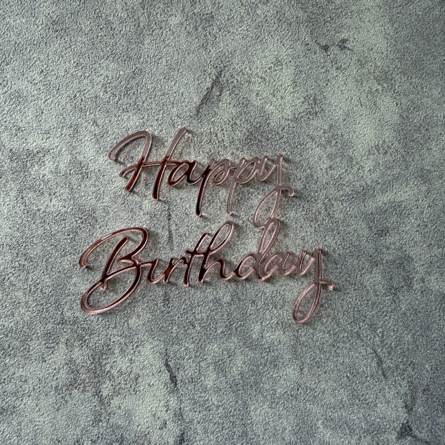 rose gold 'Happy Birthday' cake topper on a textured gray background