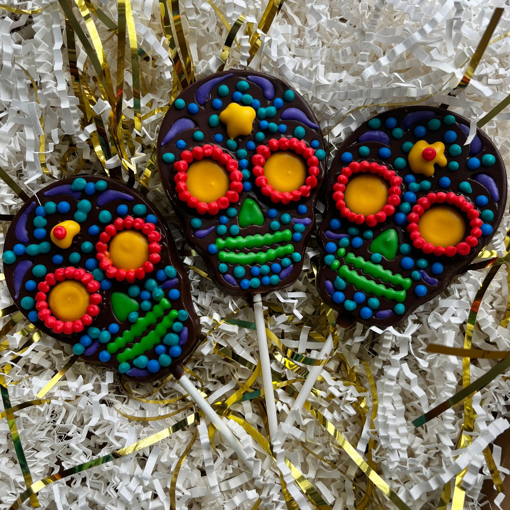 Colorful skull-shaped cookies on sticks with decorative elements on a white and gold textured background.
