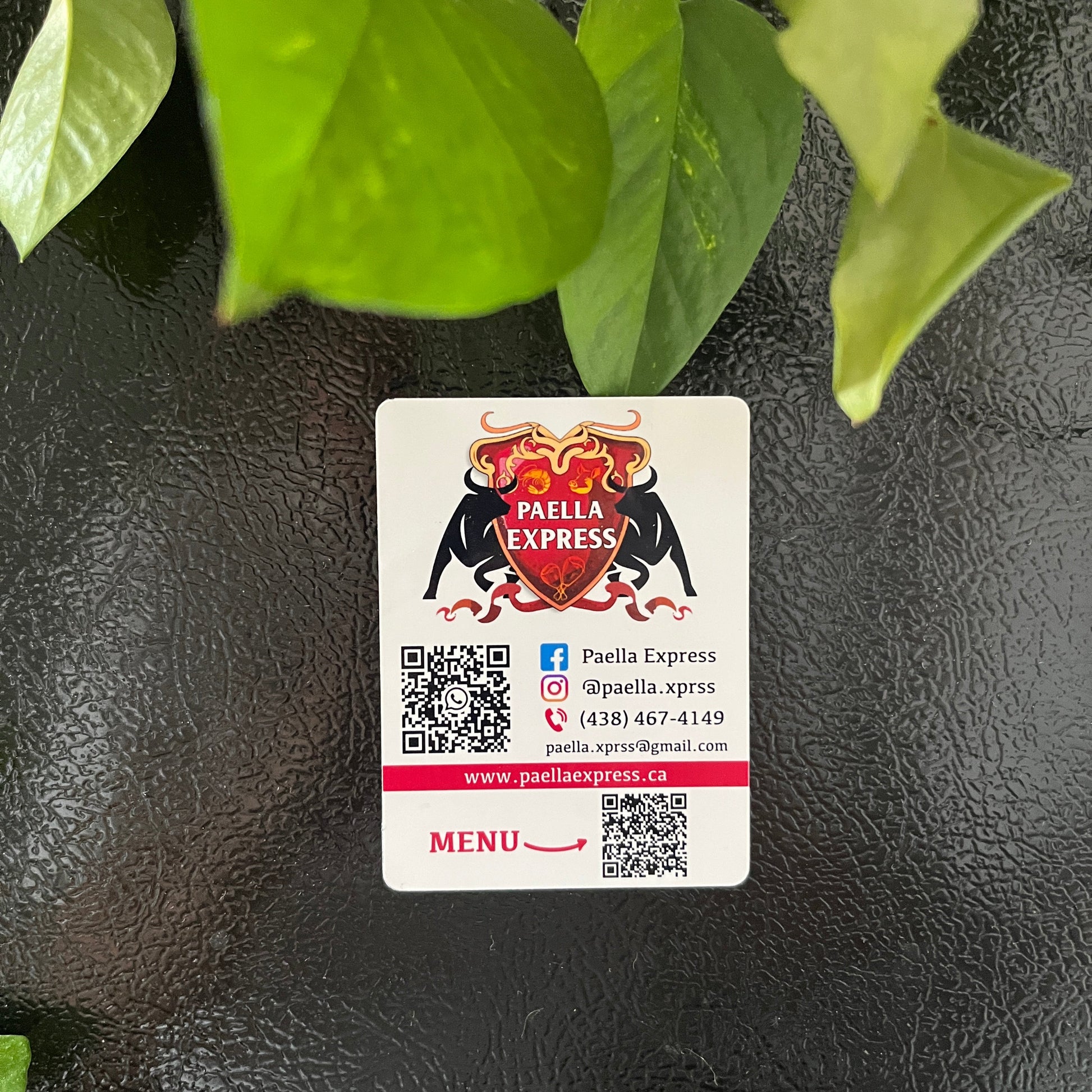 Magnet with a logo and QR code on a textured black surface with green leaves.