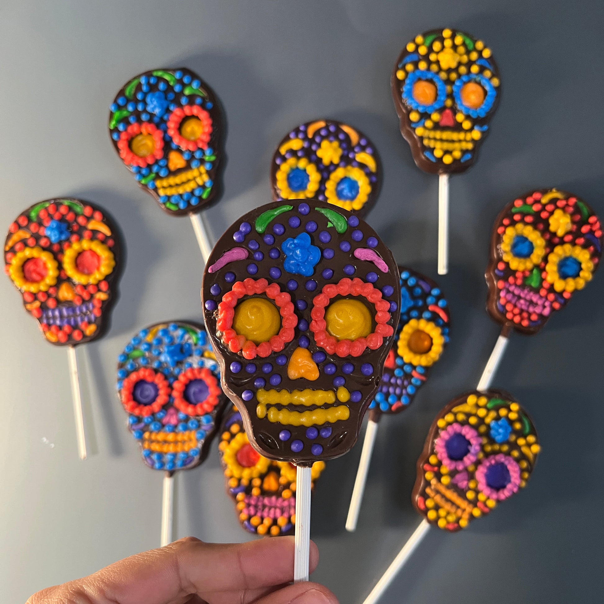 Colorful sugar skull cookies on sticks with a hand holding one, on a gray background.