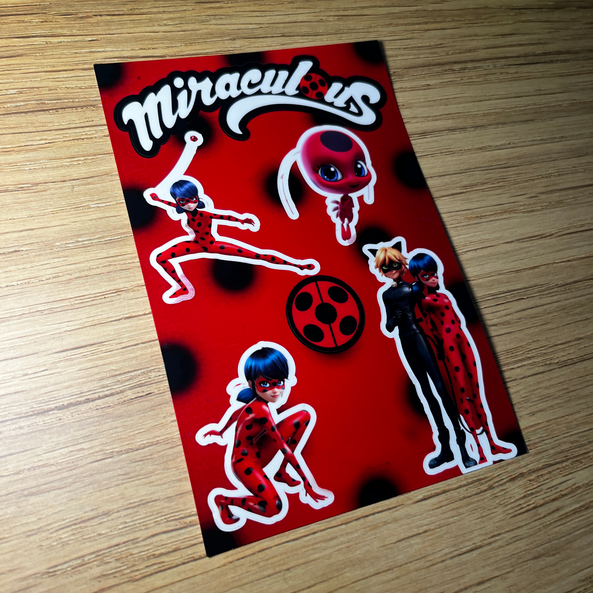 Sticker with 'Miraculous' characters on a red background, placed on a wooden surface.