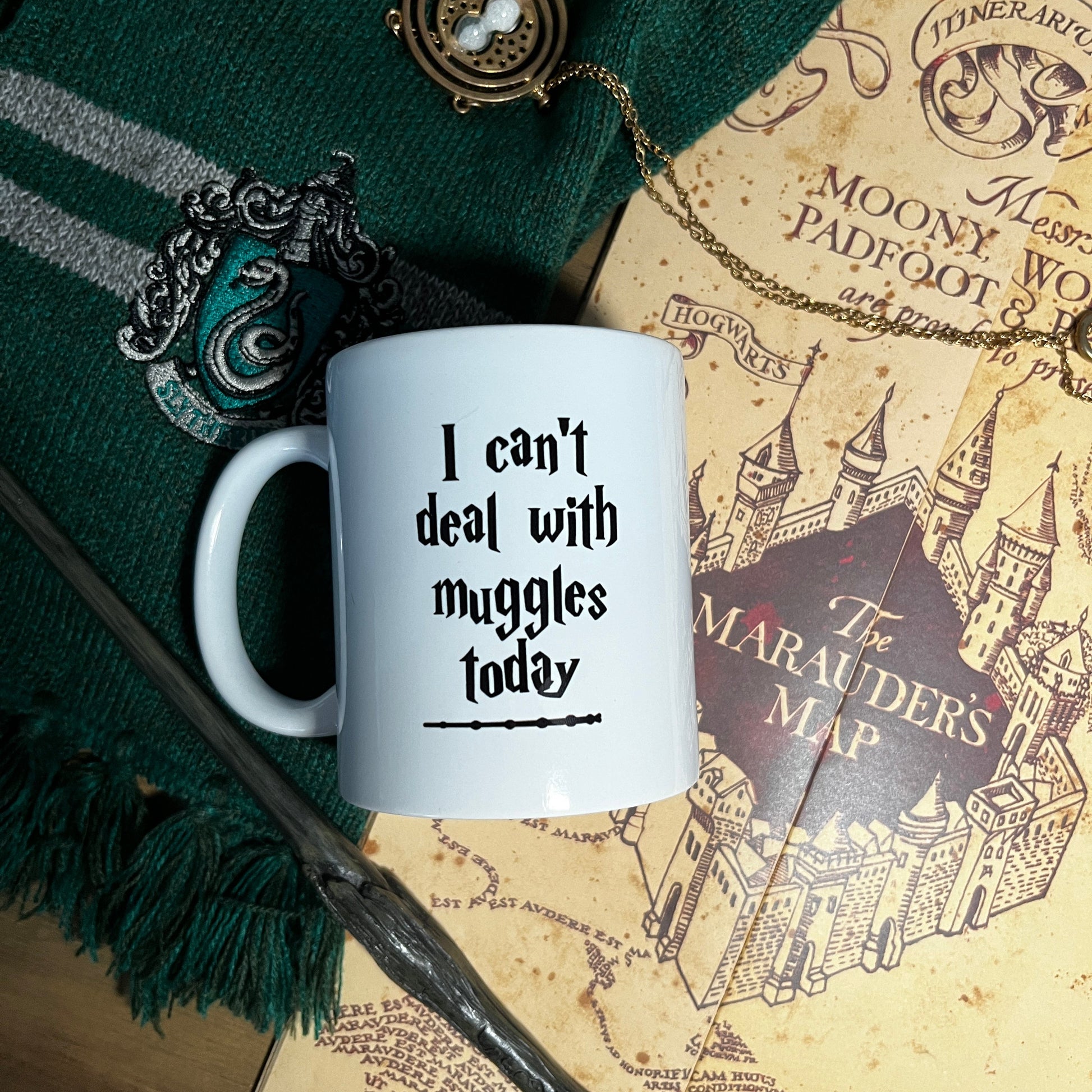 White mug with text 'I can't deal with muggles today' on a green fabric background with a wand and map.