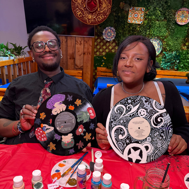 Two people holding vinyl record art pieces with a table of paint supplies in the background.