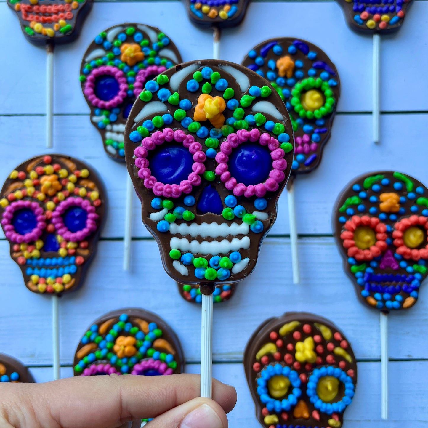 Colorful sugar skull cookies on sticks with a hand holding one, against a light blue background.